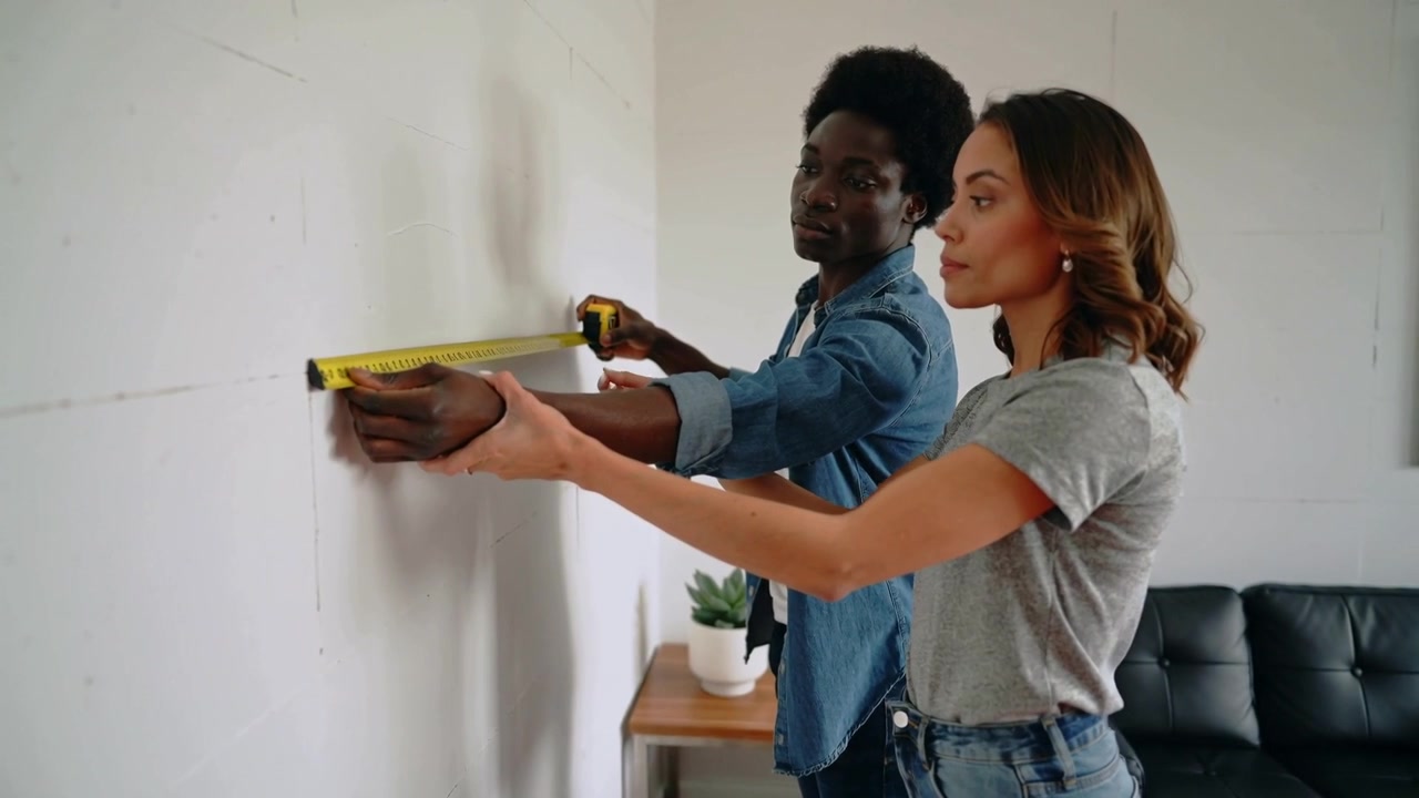 Positioning near wall, couple holding yellow tape, measuring and aligning marks at home for project
