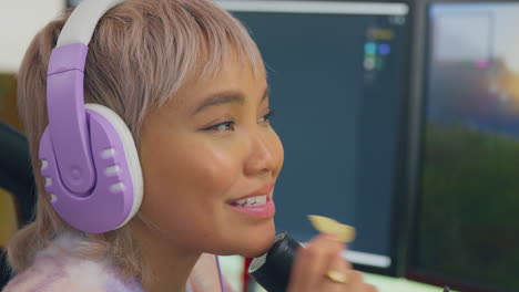 woman eating snacks whilst gaming at home sitting at desk wearing headphones for live stream