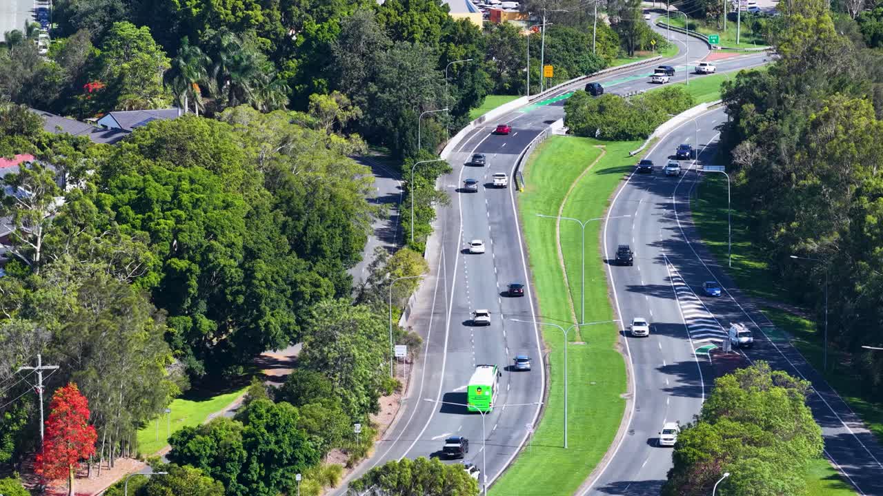 Static aerial shot of cars driving along sunny, tree-lined divided highway through suburban greenery