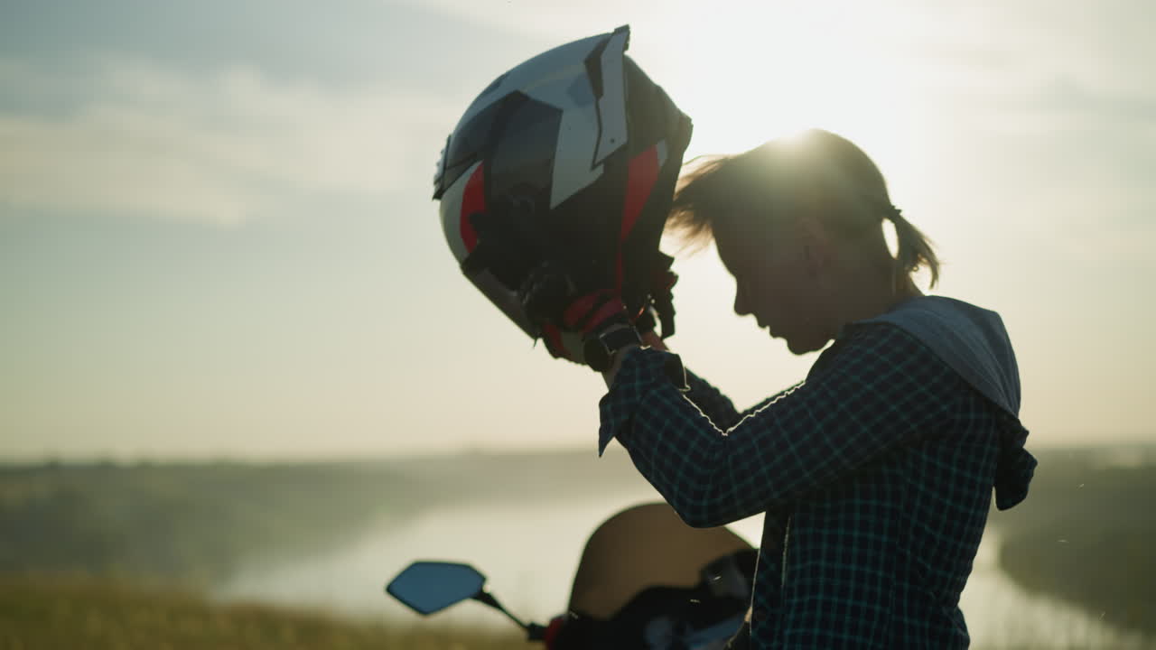 una mujer joven, en un campo cubierto de hierba, se quita el casco y ajusta su cabello mientras la brillante luz del sol se refleja en su figura, se para junto a una bicicleta