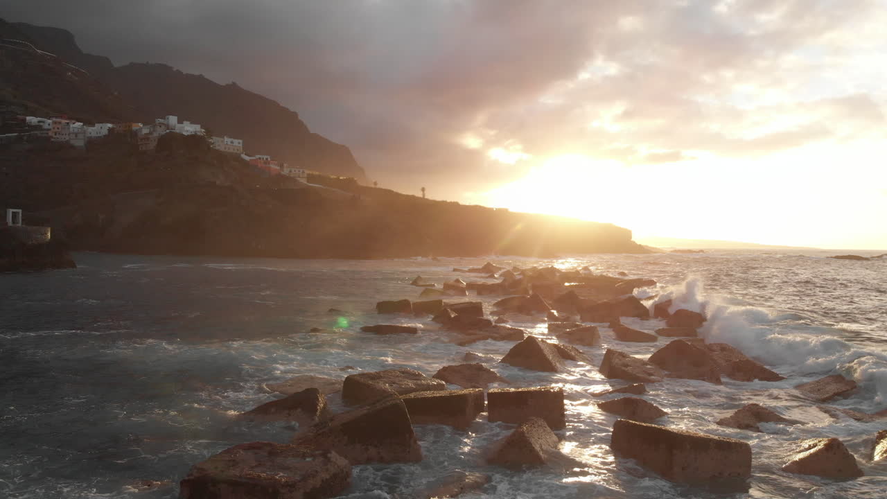 Aerial view moving fowards, Steep volcanic shore at sunset, reefs of volcanic lava, stormy ocean, white foam from the giant waves that hit rocks. Garachico, Tenerife, Canary Islands.