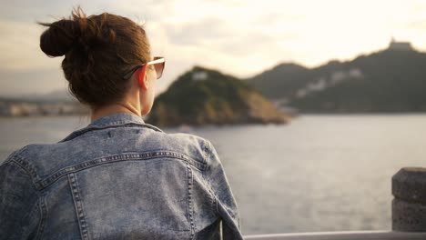 Girl-looking-out-over-harbour-coastline-during-sunset-in-San-Sebastian-Spain-Europe,-boats-on-the-water-in-a-light-breeze-in-the-warm-sun,-handheld-shot