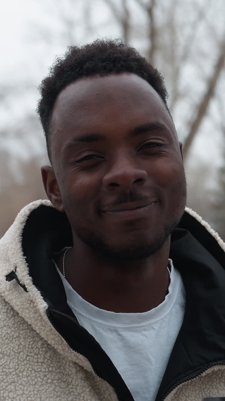 hombre sonriente con chaqueta de invierno de pie al aire libre en un entorno nevado con árboles desnudos en el fondo, su expresión cálida contrasta con la atmósfera fría