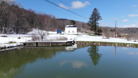 Aerial-Drone-Footage-Flying-Low-over-a-Lake-Towards-an-Old,-White,-House-on-the-Edge-of-a-Forest,-Covered-in-Snow