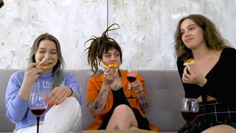 happy woman with tattoos and dreadlocks eating pizza and drinking wine while talking with friends at home