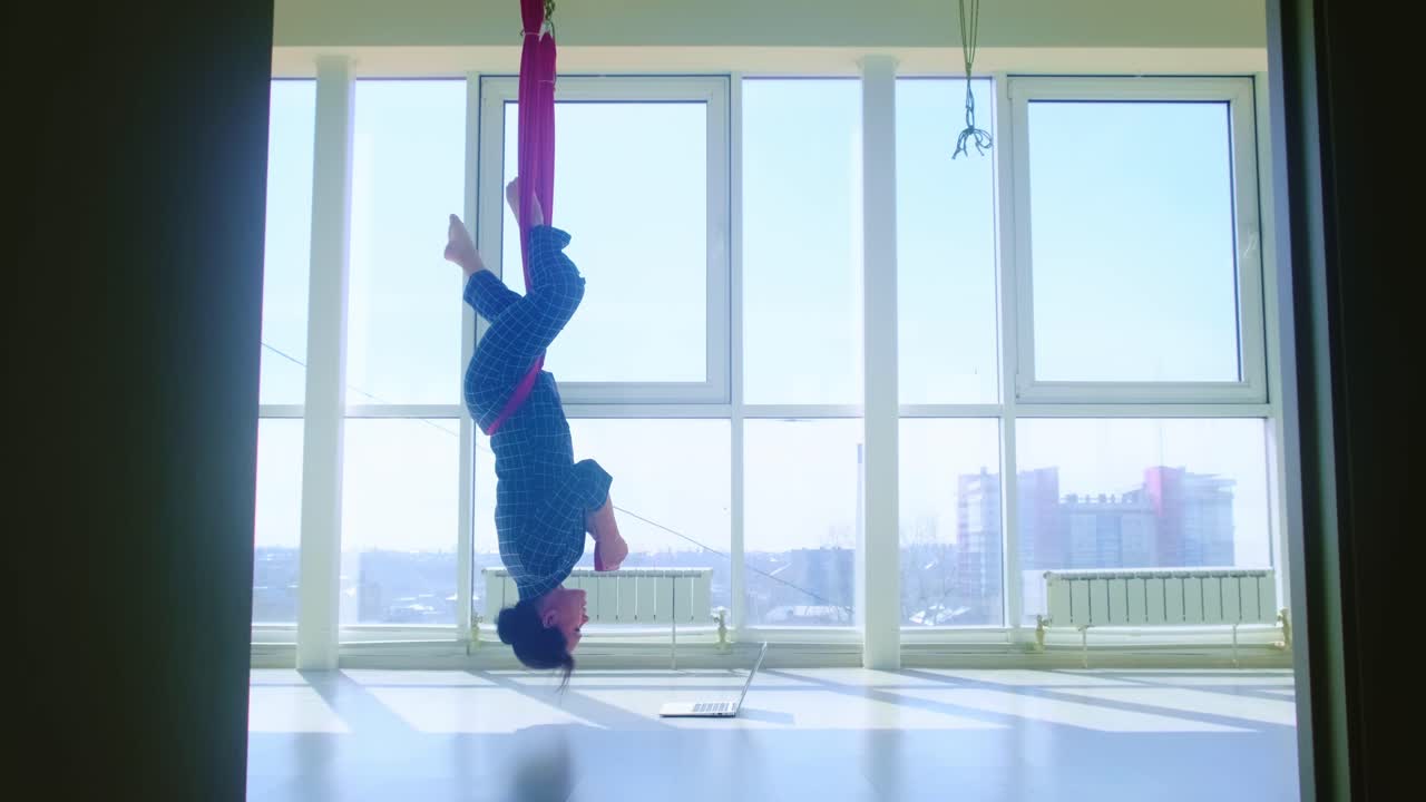 Aerial Yoga: An Individual Practicing Upside-Down Pose, Focused on a Laptop, in a Bright, Spacious Room with Large Windows