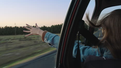 young-woman-in-car-holding-hand-out-window-feeling-wind-blowing-through-fingers-driving-in-countryside-on-road-trip-travelling-for-summer-vacation-enjoying-freedom-on-the-road-at-sunset