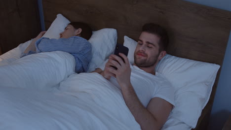 Joven-Caucásico-Sonriente-Acostado-En-La-Cama-Cerca-De-Su-Joven-Esposa-Y-Charlando-En-El-Teléfono-Inteligente-A-Altas-Horas-De-La-Noche