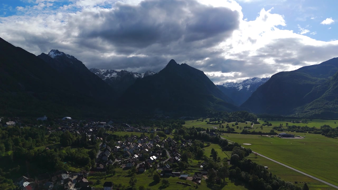 Dramatiske stormskyer svæver over sneklædte Julian Alper-toppe nær Bovec, Slovenien, grønne dalhuse ligger tæt ved en græslandingsbane omgivet af tætte skovklædte skråninger. Bredt luftfoto taget med drone