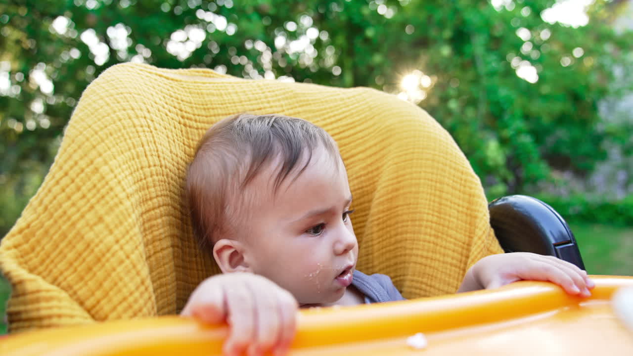 Little toddler boy sitting in a yellow feeding chair outdoors. Loving mom gives porridge to her son from spoon. Close up.