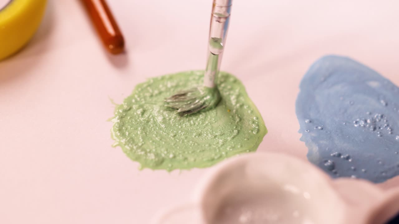 Paintbrush stirs green acrylic paint beside blue paint on pastel surface, soft studio lighting