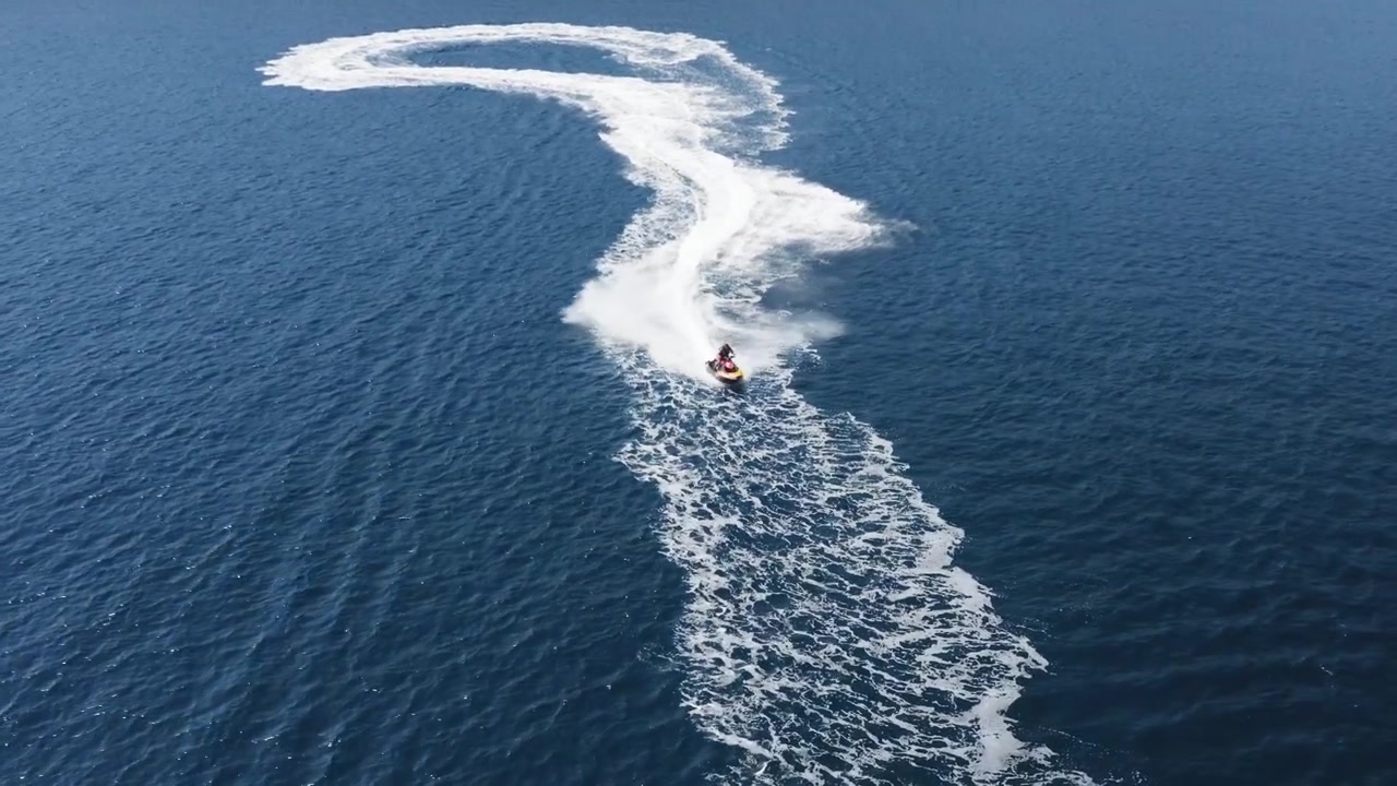 Ujęcie z drona z lotu ptaka przedstawiające osobę na skuterze wodnym, płynącą szybko. Widoczna jest linia brzegowa, ocean i fale rozbijające się od kilwateru skutera wodnego.