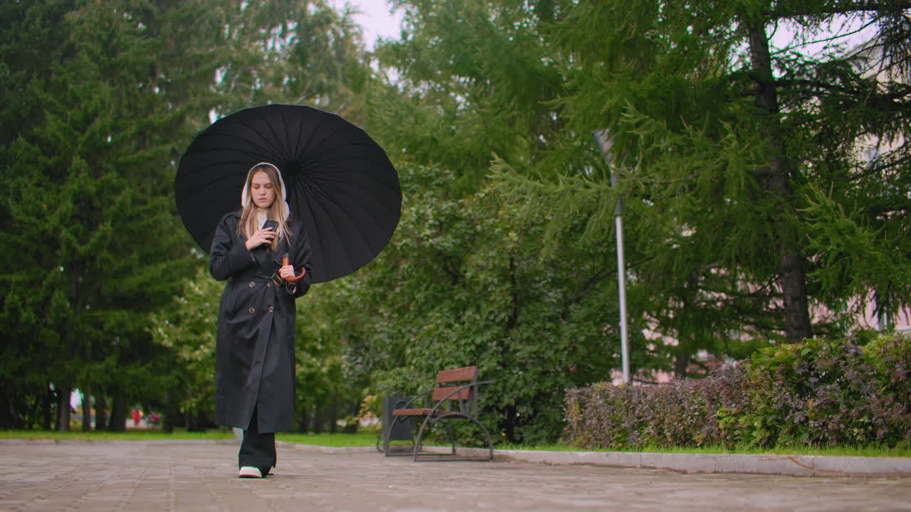 Maiden walking in park holding umbrella and talking on phone while wearing black coat and hood on cloudy rainy day, surrounded by trees, benches, and greenery lifestyle communication atmosphere