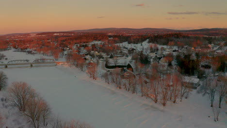 Winter-aerial-view-of-a-scenic-small-town-along-a-frozen-snow-covered-river-at-sunset