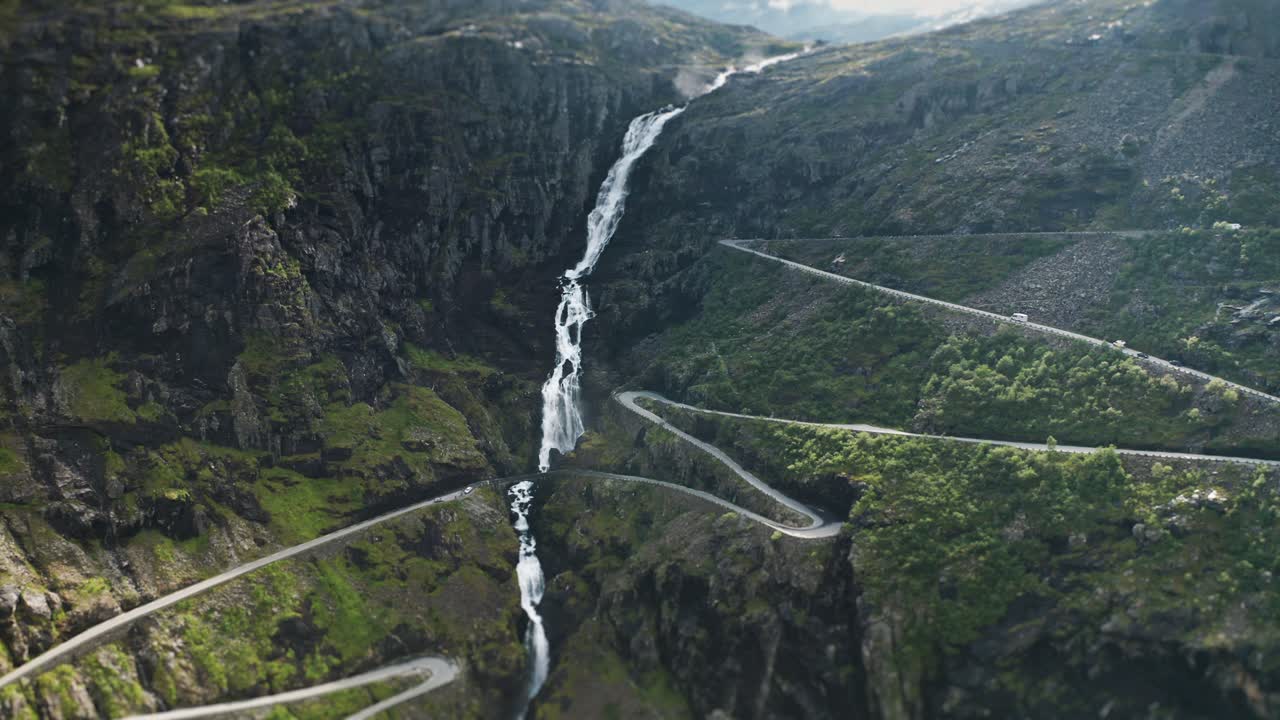 The scenic Trollstigen road, known for its sharp bends, passes beside a dramatic waterfall flowing through cliffs in the tilt-shift video.