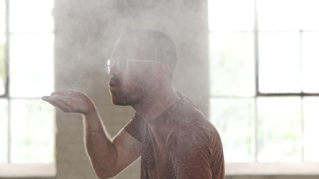 Man in an industrial warehouse wearing white glasses and moving his hand across the screen blowing flour into the air into a cloud.