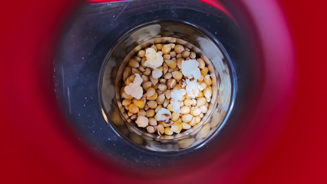 Eine Nahaufnahme von oben in eine rote Popcornmaschine, die zeigt, wie Maiskörner zu platzen beginnen. Hochwertiges Lifestyle-Filmmaterial für Kino, Snacks oder Social-Media-Inhalte für den Heimkinoabend.