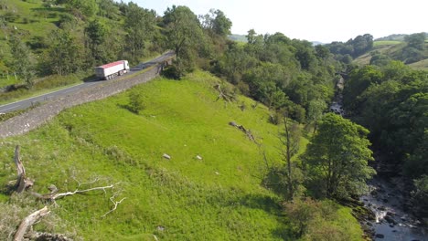 Drone-footage-reversing-in-a-shallow-tree-and-grass-lined-valley-over-a-river-and-river-bed-on-a-sunny-summer-day-in-rural-Yorkshire,-UK