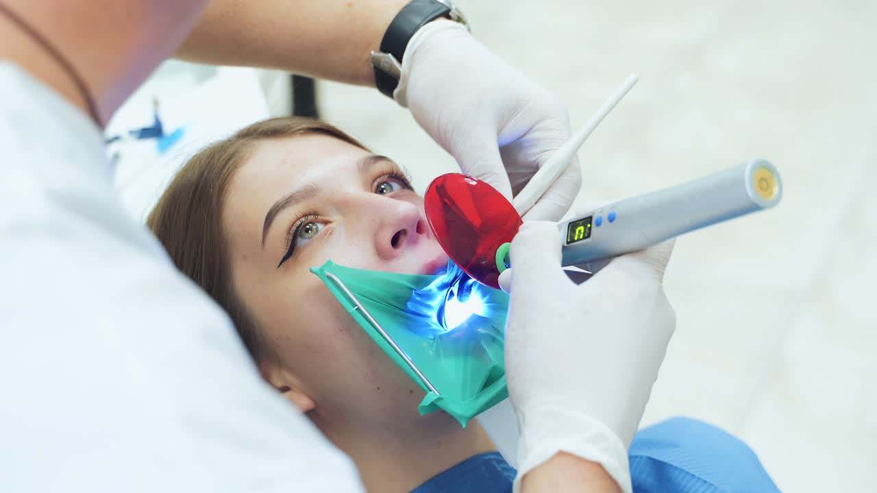 A dentist in white latex gloves uses an ultraviolet light tool to treat a tooth. A girl treats teeth for caries. Visit to the dentist