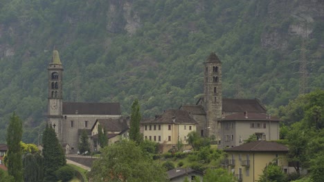 Iglesia-Católica-Y-Torres-En-Un-Pequeño-Pueblo-Rodeado-De-Un-Denso-Bosque-De-Pinos-Verdes,-Ladera-Verde-En-El-Fondo-Durante-El-Día,-Alpes-Suizos