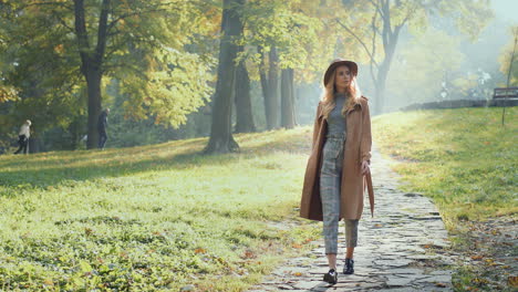 vista completa de una mujer caucásica con cabello rubio usando un abrigo y sombrero caminando en el parque por la mañana en otoño
