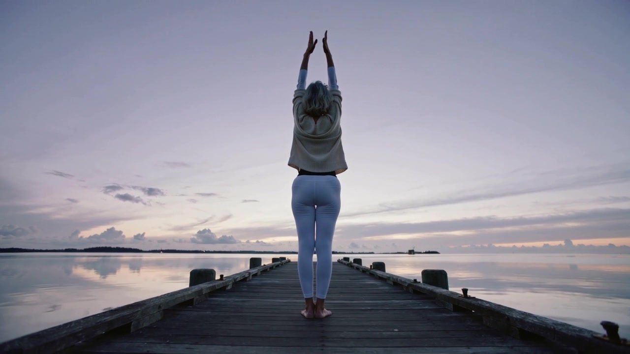 Luce dell'alba che ispira una donna che alza le braccia alla fine del molo, con un top beige e leggings bianchi
