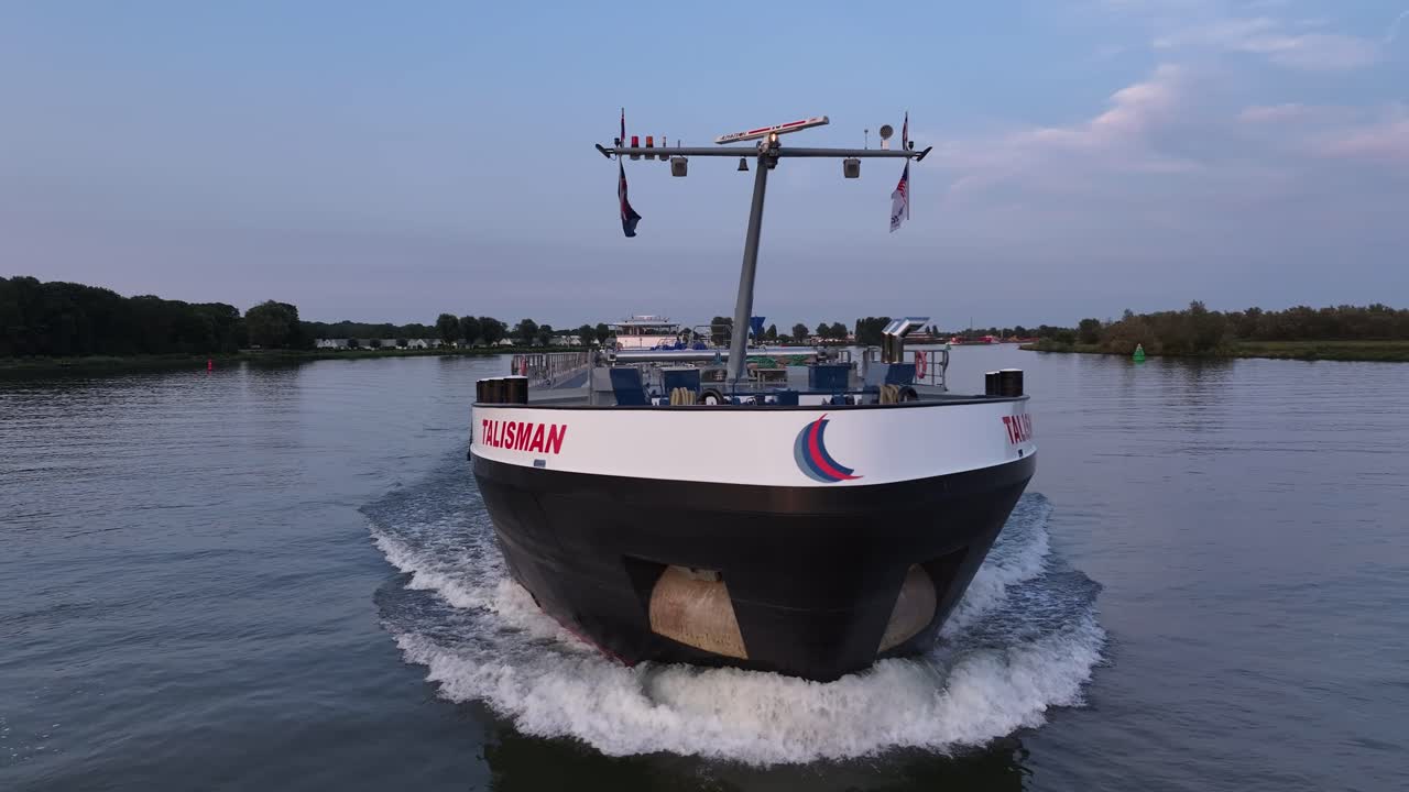 A cargo ship named Talisman cruises along a calm river during a clear day at dusk