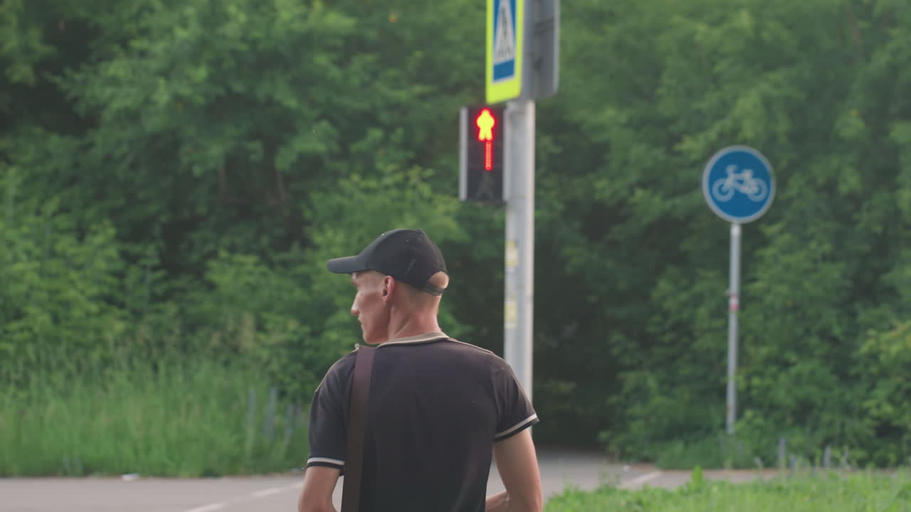 Urban Pedestrian Anticipation, Man Awaits At Crossing Light, Person Waits With Slight Movement At City Intersection, Man With Cap Pauses At Traffic Signal Amid Treelined Streets In City