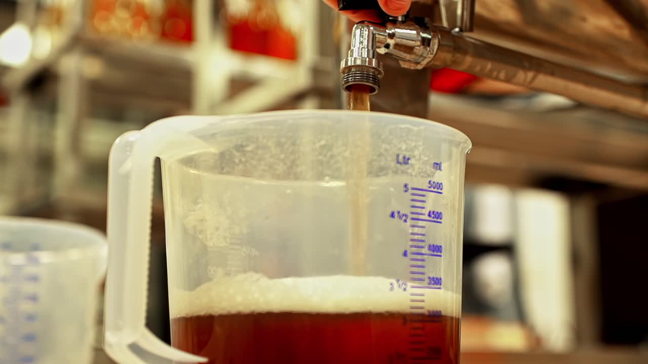A close-up shows fermented kombucha tea being poured from a tap into a large measuring pitcher during the commercial brewing and bottling process, highlighting the healthy probiotic drink