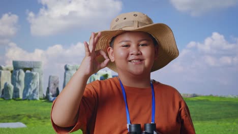 asian boy with a hat showing okay gesture after looking through the binoculars. boy researcher examines something while traveling in stonehenge, travel tourism adventure concept, close up
