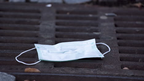 a simple green protective covid-19 face mask lying thrown out on a rectangular rusty sewerage hatch amongst dry leaves, a person picking it up carefully, static close up 4k shot