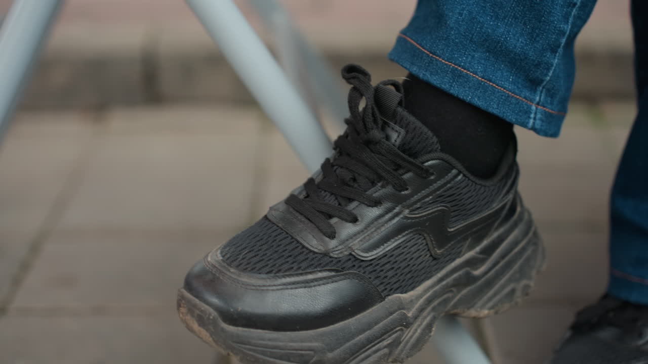 Sneaker nere su pavimentazione piastrellata, piede di un pendolare che batte sotto una sedia di metallo, polsino di jeans visibile, scarpa da ginnastica con lacci, suola usurata e segni di graffi, scena di attesa informale che suggerisce una pausa durante una camminata