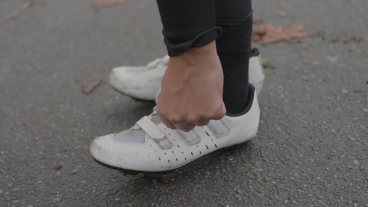 Una mano regola una scarpa da ciclismo bianca sull'asfalto all'aperto durante il giorno.