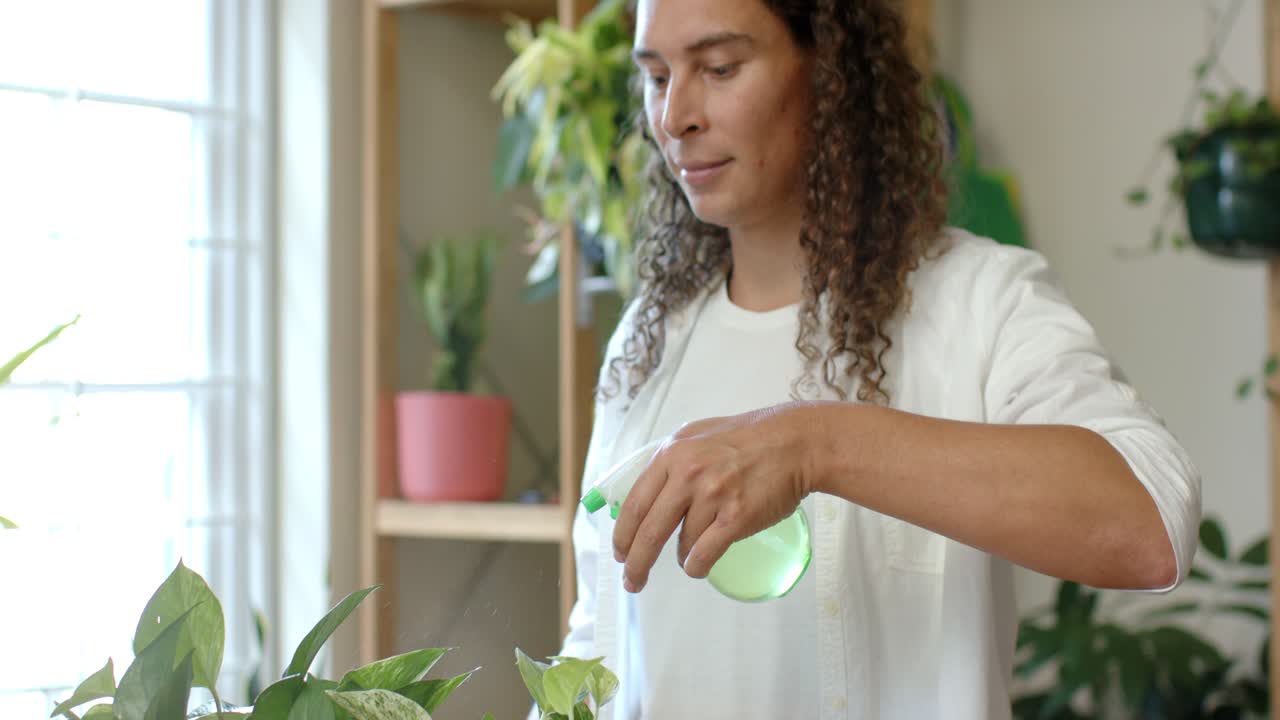 Nonbinary adult noticing dust misting pothos leaves with green mister by window, checking leaves