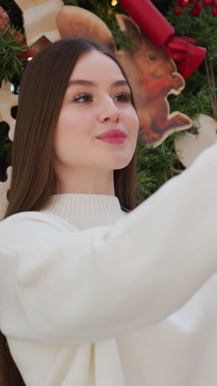 Young woman in white sweater smiling while taking selfie in front of Christmas tree in mall with blurred people in restaurant background and bokeh lights