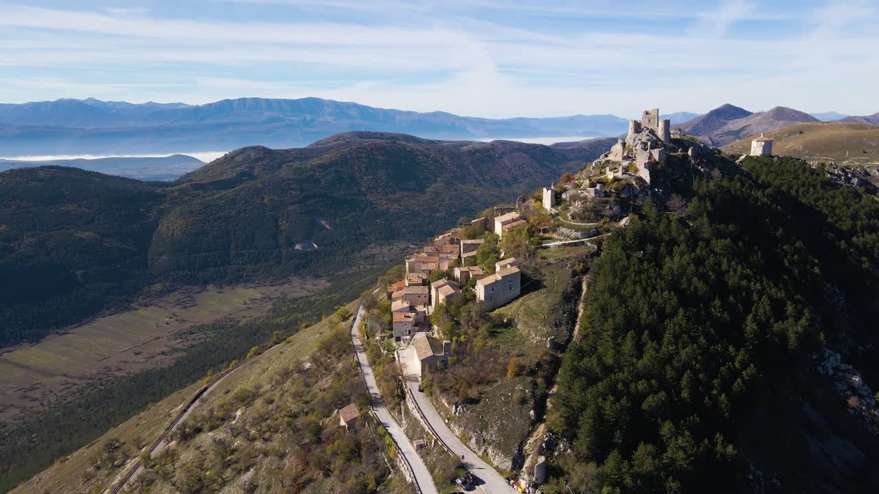 Ripresa aerea con drone della fortezza medievale e del borgo di Rocca Calascio su una cima rocciosa con una spettacolare nebbia che si muove nelle valli, campi terrazzati e un suggestivo paesaggio montano oltre.