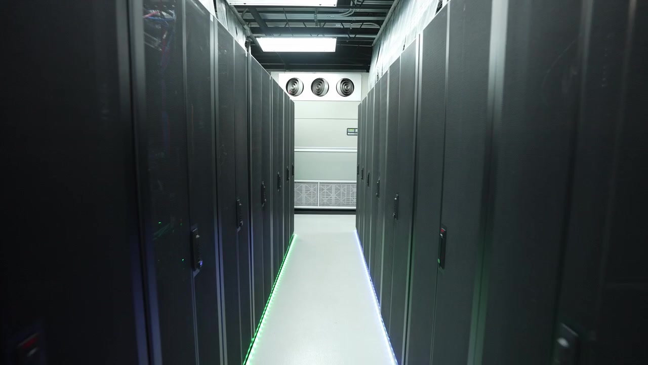 Gimbal-optagelse fremad ned ad en mørk datacentegang mellem symmetriske sorte serverrackskabe med grøn LED-gulvbelysning og industrielle køleventilatorer på endevæggen