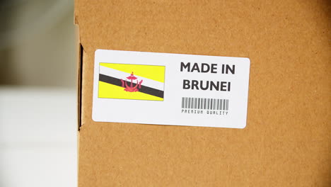 manos aplicando la etiqueta de la bandera de brunei en una caja de cartón de envío con productos