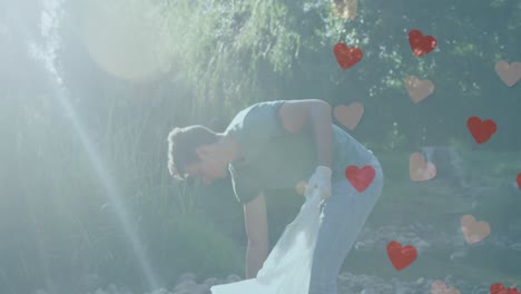 animation of hearts moving over caucasian man picking up rubbish on sunny day in countryside