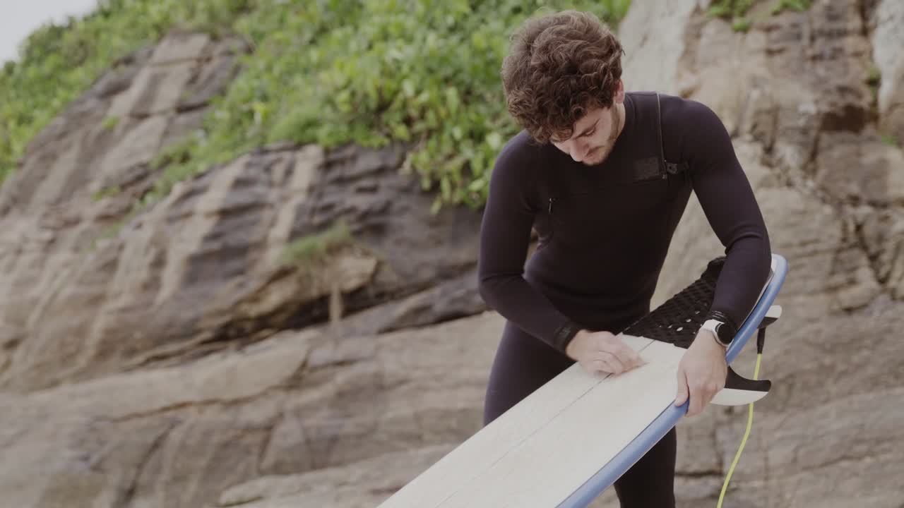 A man in a wetsuit waxing his surfboard on the beach