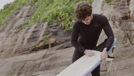 a man in a wetsuit waxing his surfboard on the beach