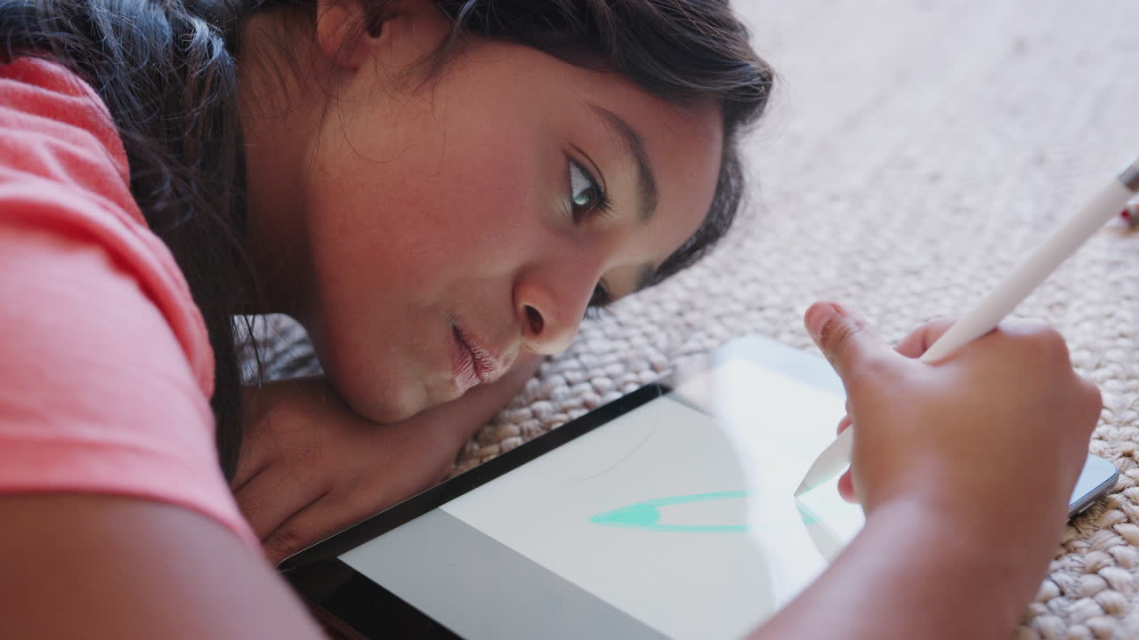 una adolescente tendida en el suelo usando una tableta y un lápiz, de cerca, vista lateral