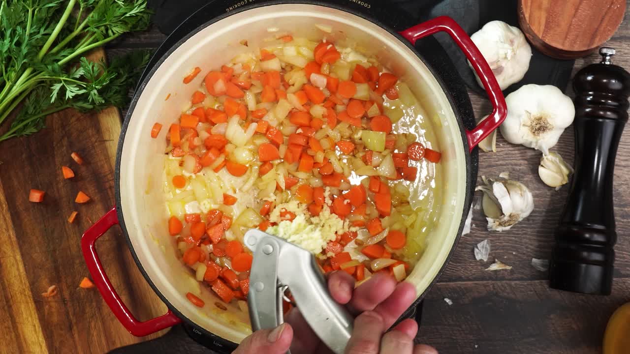 Cooking Italian Wedding Soup and adding in minced garlic using a garlic press to the red stock pot. Using a wooden spoon to stir the soup while its simmering. Adding garlic while cooking soup.