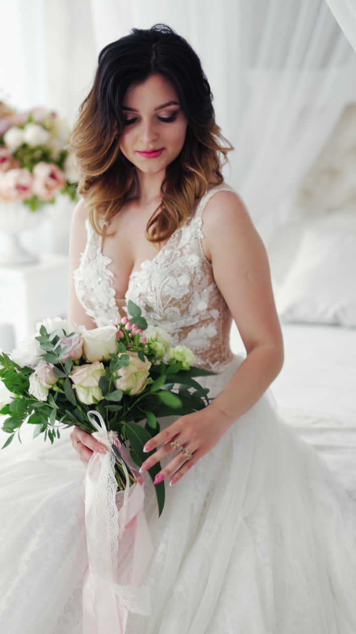 Video shot of a young bride in a wedding dress on the background of a bed. Attractive model with modern hairstyle in white dress is posing to camera inside the light room. Vertical video