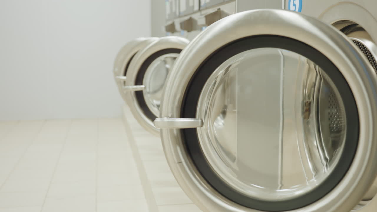 Dim room with industrial drying machines in row, doors open as light turns on, revealing metallic circular frames of equipment, modern laundromat interior, quiet atmosphere