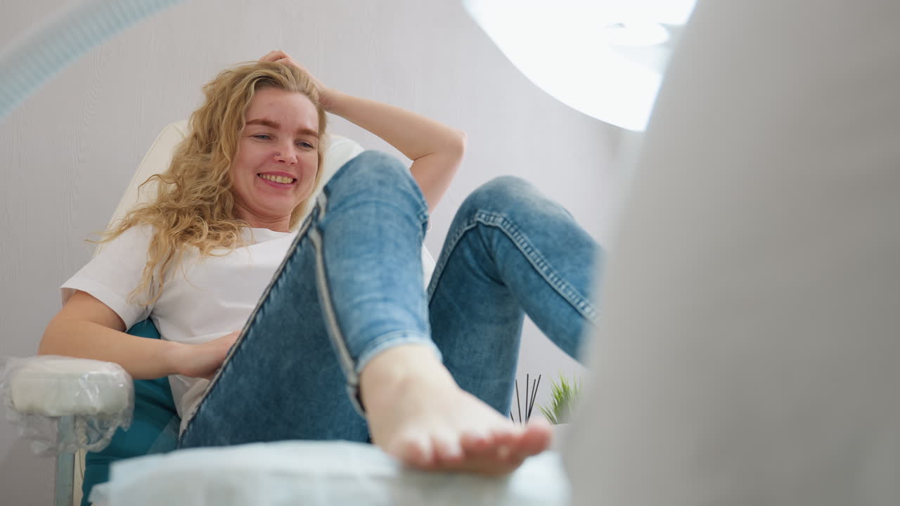 Blond-haired client in white shirt and denim jeans laughs joyfully while reclining during pedicure session. Soft lighting and modern salon setup with visible lamp highlight cheerful environment