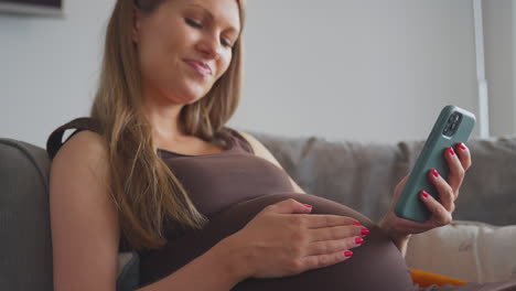 Mujer-Embarazada-Con-Teléfono-Móvil-Relajándose-En-El-Sofá-De-Casa-Sosteniendo-El-Estómago