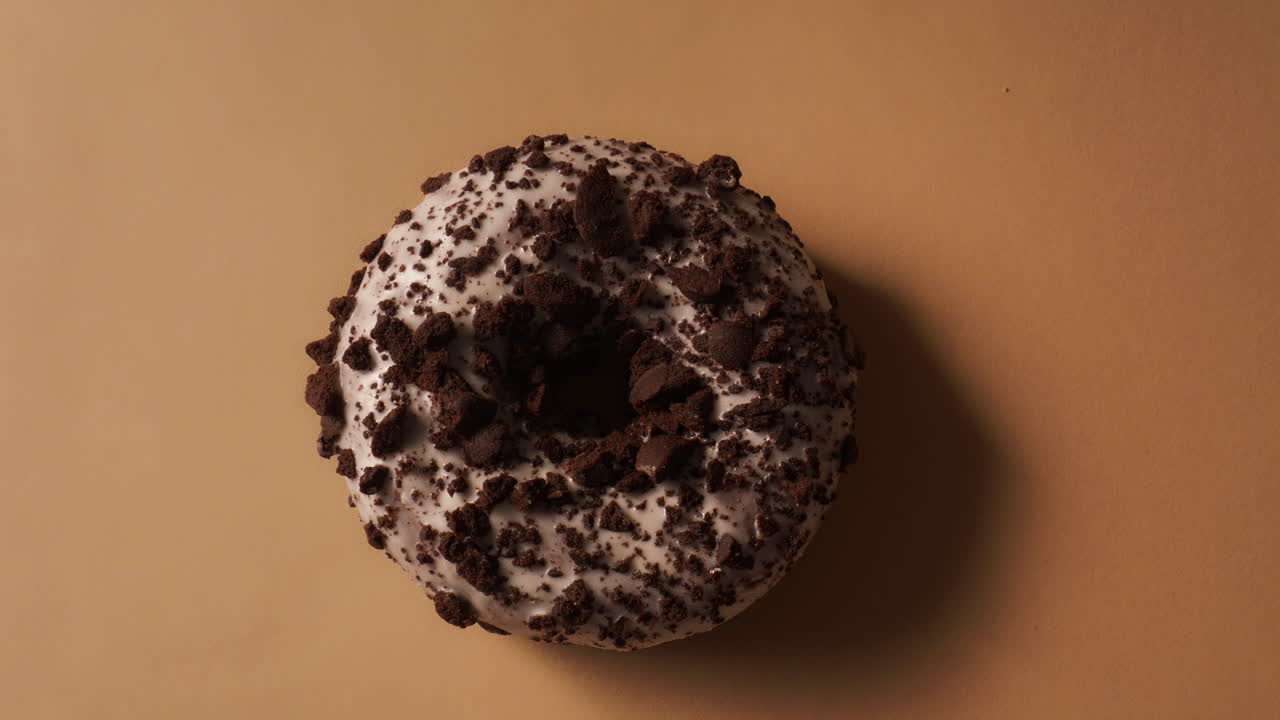 Overhead shot of hand placing chocolate donut with white icing and dark cookie crumbs onto beige surface. Rough crumb texture and glossy glaze visible in detail. Clean bakery style setup