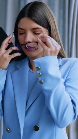 Serious beautiful young woman puts on glasses in office. Lady looks at her phone and then takes off her glasses and looks at window. Close up. Vertical video