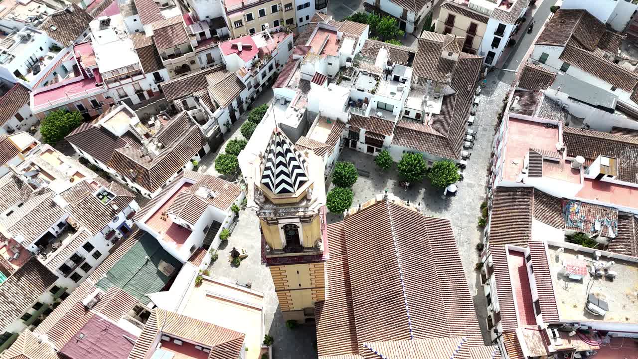 Aerial drone shot of historic church with patterned tower surrounded by narrow streets, whitewashed houses, tiled rooftops, and small plaza with trees in old town area of Ronda, Spain